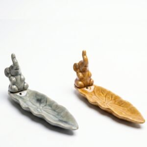 Porta sahumerios de cerámica con forma de hoja y figura de elefante con la trompa hacia arriba, colores gris azulado y ocre esmaltado, 21x6 cm.